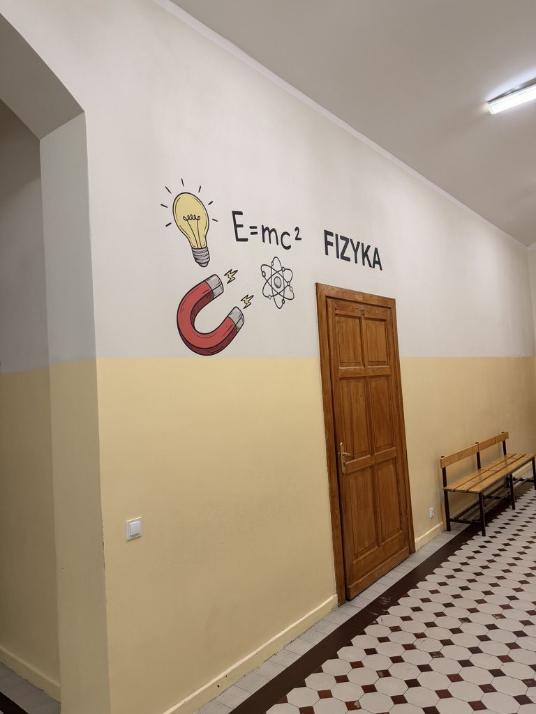 Napis FIZYKA z żarówką, wzorem E=mc² i magnesem nad drzwiami sali lekcyjnej