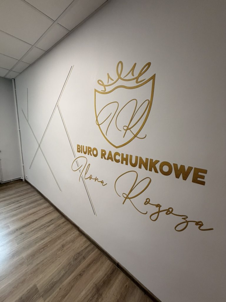 Zbliżenie na złote logo, nazwę firmy i kaligraficzny podpis Ilona Rogoza wydrukowane na ścianie