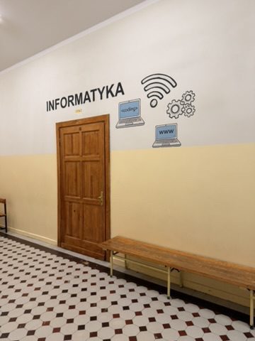 Napis INFORMATYKA z ikonami Wi-Fi, laptopów i trybików nad drzwiami sali komputerowej