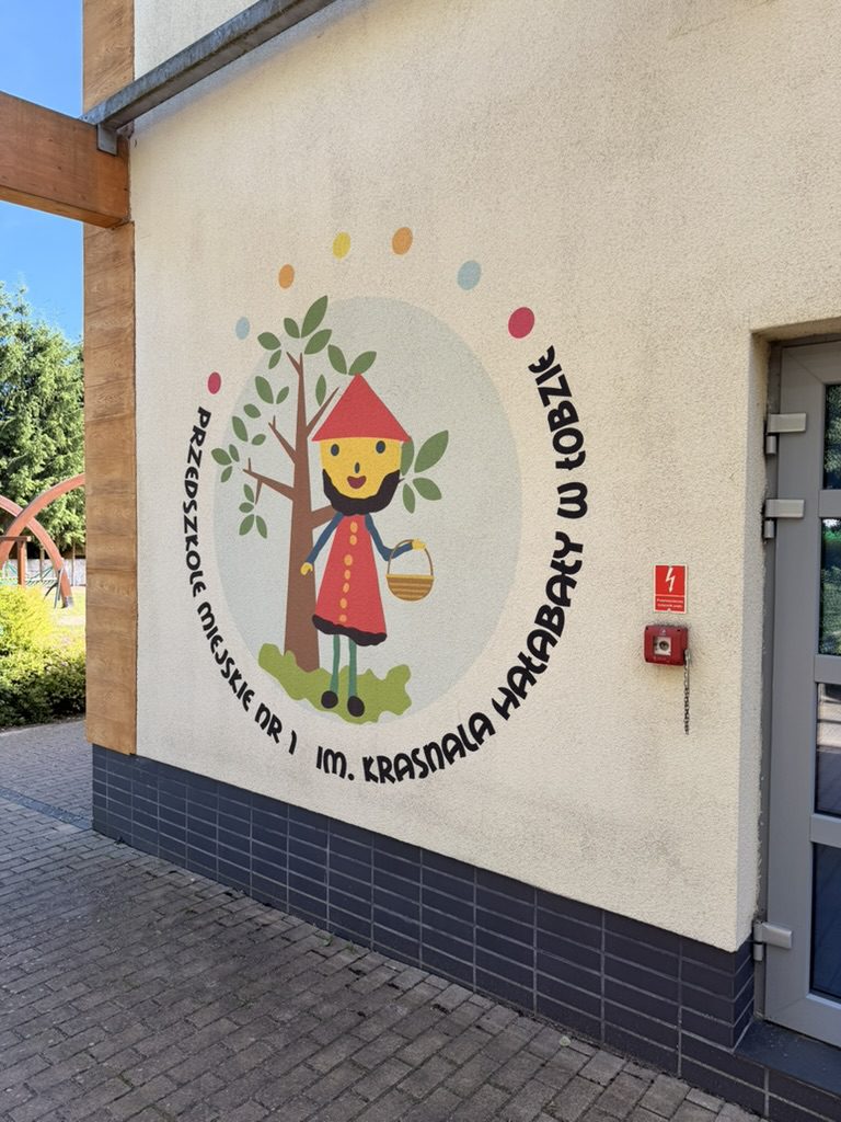 Okrągły mural z Krasnalem Hałabałą, drzewem i nazwą przedszkola wydrukowany na elewacji budynku